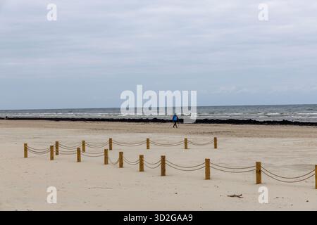 Weite malerische Aussicht auf einen riesigen Sandstrand mit einer dekorativen Seilbarriere auf Holzpfosten, eine entfernte Person und eine dunkle Algenlinie nahe dem Meer unter einem Stockfoto
