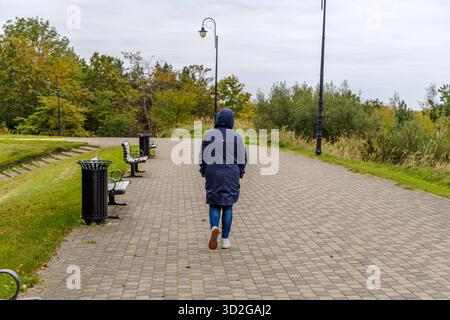 Ein Rückblick auf eine Frau in einem dunkelblauen Mantel, die an einem bewölkten Tag einen breiten, gepflasterten Weg entlang geht, der von Straßenlaternen und schwarzen Parkbänken gesäumt ist. Stockfoto