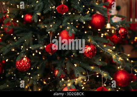 Detaillierte Nahaufnahme eines lebendigen Weihnachtsbaums mit dunkelgrünen Nadeln, leuchtenden gelben Lichtern und verschiedenen roten Weihnachtsschmuck. Stockfoto