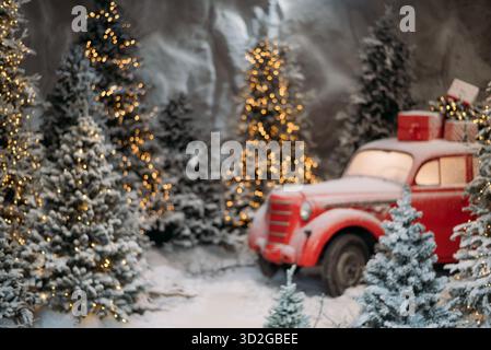 Verschwommenes Foto eines roten Oldtimers mit Geschenken auf der Oberseite, geparkt in einer verschneiten Winterwaldszene mit leuchtenden, dekorierten Weihnachtsbäumen. Stockfoto
