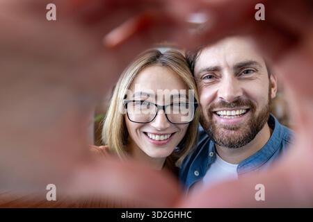 Glückliches, vielseitiges Paar, das ein Herz mit den Händen um das Gesicht zaubert und die Kamera in einem warmen, ehrlichen Innenporträt anlächelt, das Liebe und Zweisamkeit ausstrahlt Stockfoto