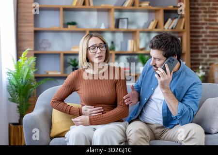 Frau mit starken Bauchschmerzen auf der Couch, hält ihren Magen, während ein besorgter Mann einen Anruf macht, wahrscheinlich sucht er medizinische Hilfe für ihren Zustand Stockfoto