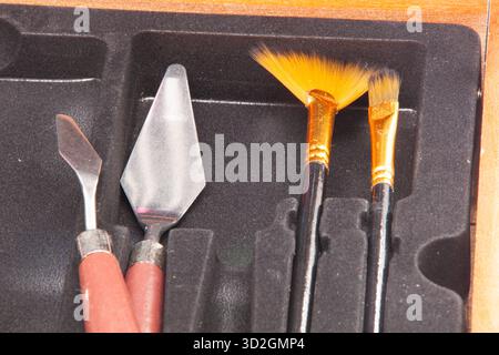 Nahaufnahme eines Satzes von Kunstwerkzeugen: Zwei Metallpalettenmesser mit roten Griffen und zwei Bürsten mit goldenen Aderendhülsen und schwarzen Griffen. Stockfoto