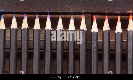 Eine Makroaufnahme von geschärften Spitzen schwarzer Bleistifte, die dicht in einer einzigen dichten Reihe angeordnet sind. Es sind Blau-, Violett-, Rot- und Orangenschattierungen sichtbar. Stockfoto