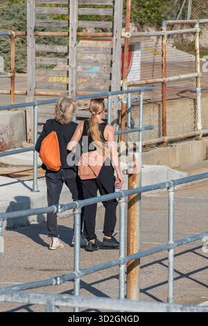 Zwei junge Frauen, die an einem Sommertag Arm in Arm auf einem Weg durch ein Industriegebiet laufen. Freundinnen, die zusammen an Geländern und Gerüsten vorbeilaufen Stockfoto