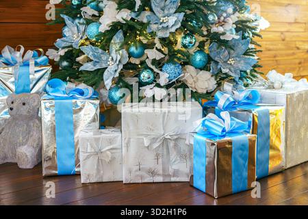Elegant gewickelte Geschenke, Geschenkboxen werden unter einem geschmückten Weihnachtsbaum platziert, der mit blauen und silbernen Ornamenten verziert ist, um fröhliche Feiertage zu schaffen Stockfoto