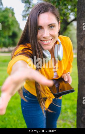 Lächelnde junge Frau draußen, die Hand nach vorne streckt und das Smartphone hält. Stockfoto