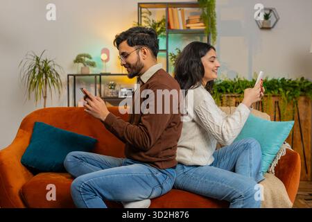 Seitenansicht eines fröhlichen, abwechslungsreichen Paares in legeren Kleidern, das sich mit Smartphones auf dem Sofa im Zimmer zu Hause zurücklehnen konnte. Eine junge indische Familie, die im Apartment auf der Couch im Handy surft Stockfoto