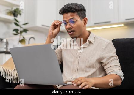 Ein indischer Mann zu Hause hat Schwierigkeiten, winzige Texte auf dem Laptop zu lesen, zieht dann eine Brille auf und sieht klare Sicht. Arabian Guy on Sofa Lächeln passt den Rahmen an, liest Bildschirmlinien ohne Belastung und setzt die Arbeit fort. Stockfoto