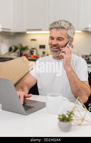 Ein Mann mittleren Alters sitzt zu Hause am Tisch, arbeitet am Laptop und spricht mit dem Kunden über das Smartphone. Reifer Mann auf Sofatypen antwortet, spricht eine ruhige Stimme verwaltet die Aufgabenliste und hält den Fokus unter Last klar und deutlich Stockfoto