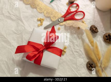 Weihnachtsgeschenk in weißes Papier gewickelt und von Bastelbedarf umgeben. Das Konzept der Materialpflege und des Recyclings. Handgefertigte Weihnachtsgeschenke Stockfoto