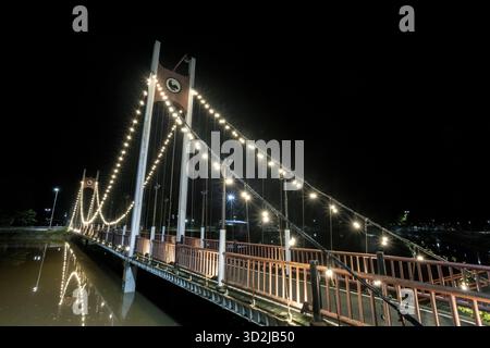 Die beleuchtete Hängebrücke leuchtet nachts hell und reflektiert das ruhige Wasser. Seine leuchtende urbane Architektur schafft Ruhe Stockfoto