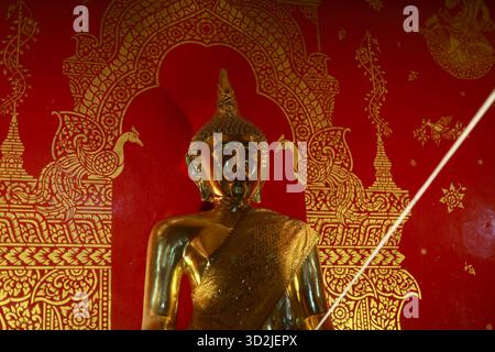 Goldene Buddha-Statue mit heiterem spirituellem Ausdruck im thailändischen Tempel. Rote Hintergrundewand mit kompliziertem goldenen Kunstwerk-Muster. Heiliges Symbol des pe Stockfoto