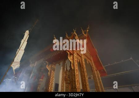 Heiliger goldener thailändischer Tempel in der Nacht, umhüllt von geheimnisvollem Rauch. Dramatische Lichtstrahlen beleuchten die traditionelle buddhistische Architektur und schaffen spirituelle Stockfoto