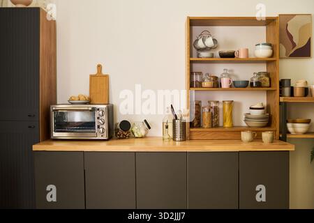 Moderne Küchenarbeitsplatte mit elektrischem Toaster, Holzschneidebrett, Glasgläsern mit Körnern und Nudeln, Keramikschalen, Tassen, Utensilien, offenen Regalen, gerahmte Wandbilder Stockfoto