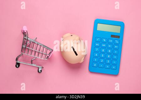 Schweinebank, Mini-Supermarkt-Trolley und Taschenrechner auf rosa Pastellhintergrund. Minimalistische Studioaufnahme. Draufsicht. Flache Lagen. Stockfoto