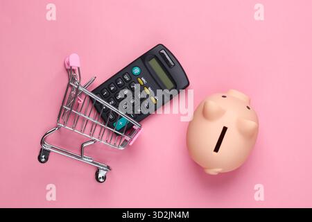 Schweinebank, Mini-Supermarkt-Trolley und Taschenrechner auf rosa Pastellhintergrund. Minimalistische Studioaufnahme. Draufsicht. Flache Lagen. Stockfoto