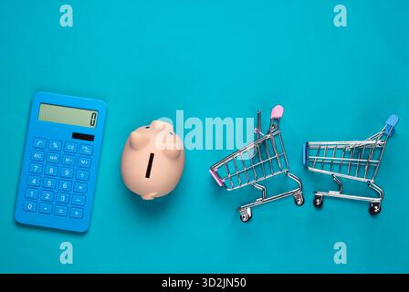 Schweinebank, Mini-Supermarkt-Trolley und Taschenrechner auf blauem Hintergrund. Minimalistische Studioaufnahme. Draufsicht. Flache Lagen. Stockfoto