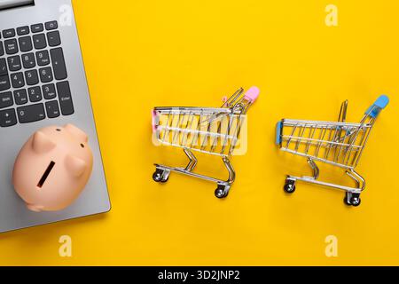 Online-Shopping. Laptop mit Piggy Bank, Supermarktwagen auf gelbem Hintergrund. Minimalistische Studioaufnahme. Draufsicht. Flache Lagen. Stockfoto