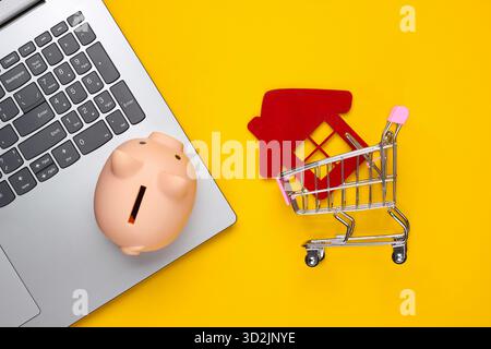 Sparen Sie für das Wohnen. Laptop, Schweinebank, Einkaufswagen mit Hausfigur, Brieftasche auf gelbem Hintergrund. Draufsicht Stockfoto