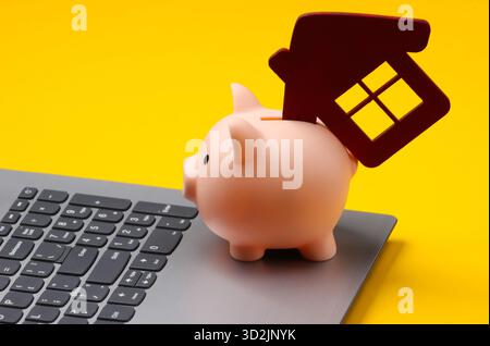 Sparen Sie für das Wohnen. Laptop, Schweinebank mit Hausfigur auf gelbem Hintergrund. Stockfoto