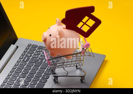 Sparen Sie für das Wohnen. Laptop, Einkaufswagen mit Schweinebank, Hausfigur auf gelbem Hintergrund. Stockfoto