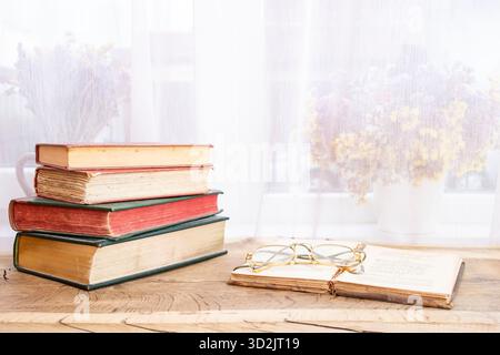 Vintage-Bücher mit festem Einband, gestapelt mit einem geöffneten Buch auf einem hellen Holztisch mit einer Brille und weißen Vorhängen Stockfoto