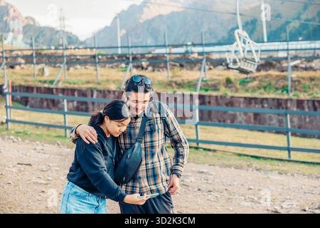 Eine junge asiatische Frau und ein asiatischer Mann mittleren Alters machen während ihres Urlaubs ein Selfie vor Bergen und einer Seilbahn. Stockfoto