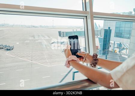 Hände einer Frau halten ein Smartphone, während sie am Flughafen wartet. Sie trägt bunte Scrunchies an ihren Handgelenken. Die Ansicht zeigt ein Flugzeug draußen. Stockfoto