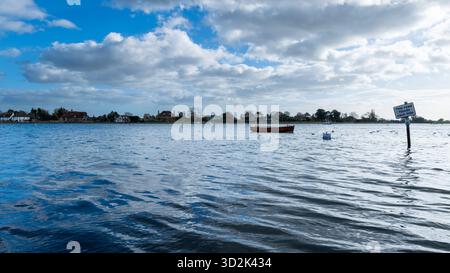 Bosham Harbour, West Sussex, England, bei Flut mit einem vertäuten Holzboot und einem überfluteten Damm-Schild unter einem hellblauen Himmel mit Wolken Stockfoto