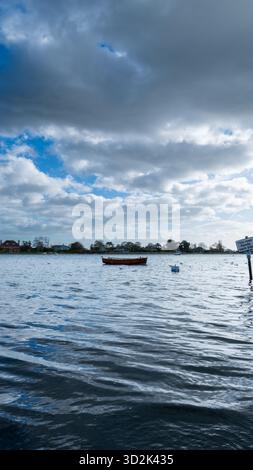 Bosham Harbour, West Sussex, England, bei Flut mit einem vertäuten Holzboot und einem überfluteten Damm-Schild unter einem hellblauen Himmel mit Wolken Stockfoto