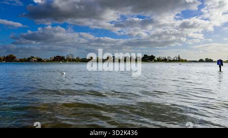 Bosham Harbour, West Sussex, England, bei Flut mit einem vertäuten Holzboot und einem überfluteten Damm-Schild unter einem hellblauen Himmel mit Wolken Stockfoto