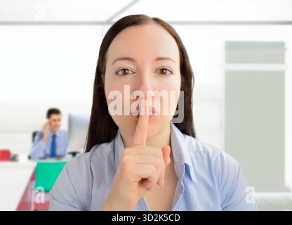 Porträt einer jungen Geschäftsfrau mit dem Finger auf den Lippen im Büro, während die Kollegin im Hintergrund arbeitet Stockfoto