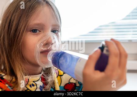 Mädchen mit Inhalator mit Spacer und Maske während der Atemtherapie zu Hause. Das Konzept des kindlichen Asthmas, der Atemstörung und der medizinischen Behandlung. Stockfoto