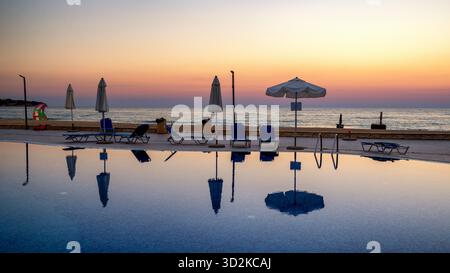 Ruhiger Sommerabend im Küstenhotel in Obzor, Bulgarien. Sonnenliegen und Sonnenschirme spiegeln sich im Swimmingpool mit Blick auf das ruhige Meer bei Sonnenuntergang. Stockfoto