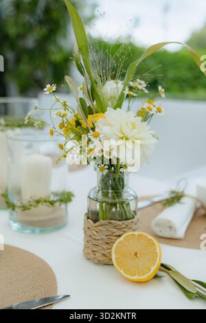 Elegante Tischeinrichtung mit einem Strauß gelber und weißer Blumen in einer Glasvase, kombiniert mit einer halben Zitrone und gewebten Akzenten. Ideal für Hochzeiten Stockfoto