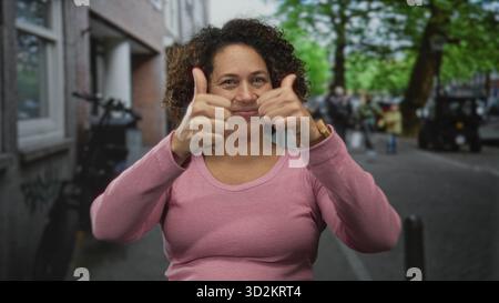 Frau in rosa Pullover gibt auf der belebten Stadtstraße, die von Bäumen und geparkten Fahrrädern gesäumt ist, zwei Daumen hoch; Optimismus. Stockfoto