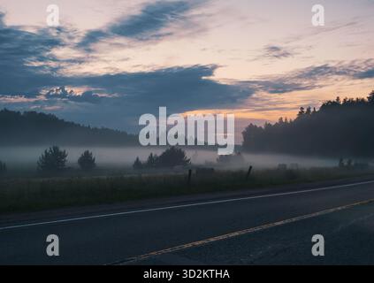 Morgens rollt Nebel über ein Feld in ländlicher Umgebung in der Nähe einer zweispurigen Straße Stockfoto