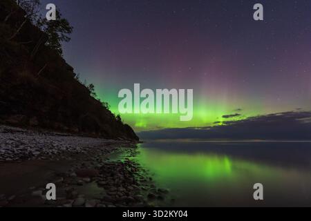 Atemberaubende grüne und violette Aurora Borealis (Nordlichter), die sich in ruhigem Wasser an der felsigen Küste Estlands spiegeln. Stockfoto