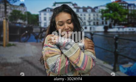 Frau umarmt ihre Schultern auf der Straße am amsterdamer Kanal mit geschlossenen Augen und einem leichten Lächeln; Selbstfürsorge-Gelassenheit. Stockfoto