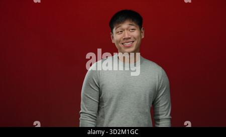 Junger chinesischer Mann, der in einem grauen Pullover vor einem isolierten roten Hintergrund lächelt und Glück und Einfachheit ausdrückt. Stockfoto