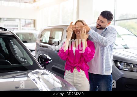 Ein verspielter Mann, der die Augen seiner Frau im Ausstellungsraum des Autos bedeckt und das überraschende Auto enthüllt Stockfoto