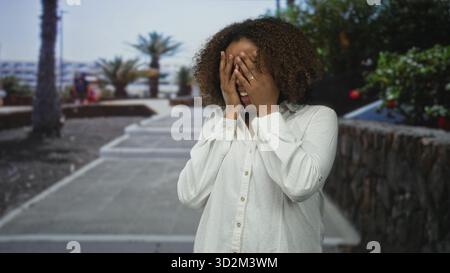 Frau, die Gesicht mit beiden Händen auf einer Straßenpromenade neben einer Steinmauer und Palmen hält, mit weißem Hemd; Überraschung. Stockfoto