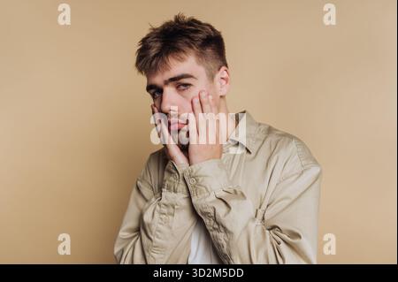 Junger Mann mit einem nachdenklichen Ausdruck, Hand auf Gesicht, beigefarbenem Hintergrund. Stockfoto