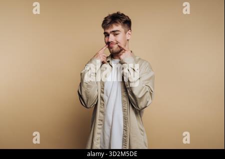 Junger Mann in lässigem Outfit lächelt und zeigt auf sein Lächeln vor beigefarbenem Hintergrund. Stockfoto
