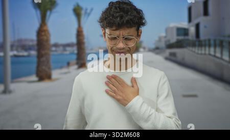 Mann Clutch Brust in weißem Pullover auf der Straße an der von Palmen gesäumten Strandpromenade am blauen Meer; Schmerzen. Stockfoto