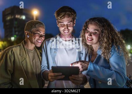 Drei lächelnde, vielfältige Freunde stehen nachts draußen und schauen sich einen gemeinsamen digitalen Tablet-Bildschirm an, während sie interagieren und Inhalte gemeinsam genießen Stockfoto