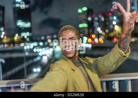 Glückliche junge schwarze Frau, die ein Selfie macht und ein Friedenszeichen zeigt, hell in die Kamera lächelt und eine pulsierende Nacht in der Stadt mit verschwommenen Lichtern genießt Stockfoto