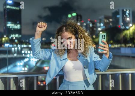 Junge Frau mit lockigem Haar, die Musik über Kopfhörer genießt, ein Smartphone hält und frei auf einer Brücke tanzt, die nachts eine geschäftige Stadt überblickt. Stockfoto