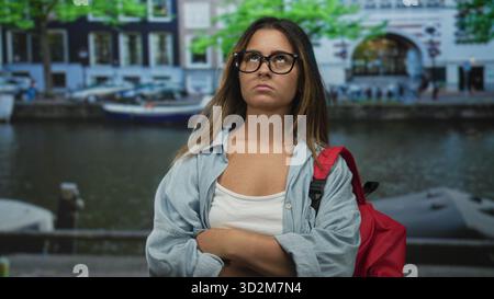 Frau mit überkreuzten Armen, Brille und rotem Rucksack auf einer Straße am amsterdamer Kanal; Frustration. Stockfoto